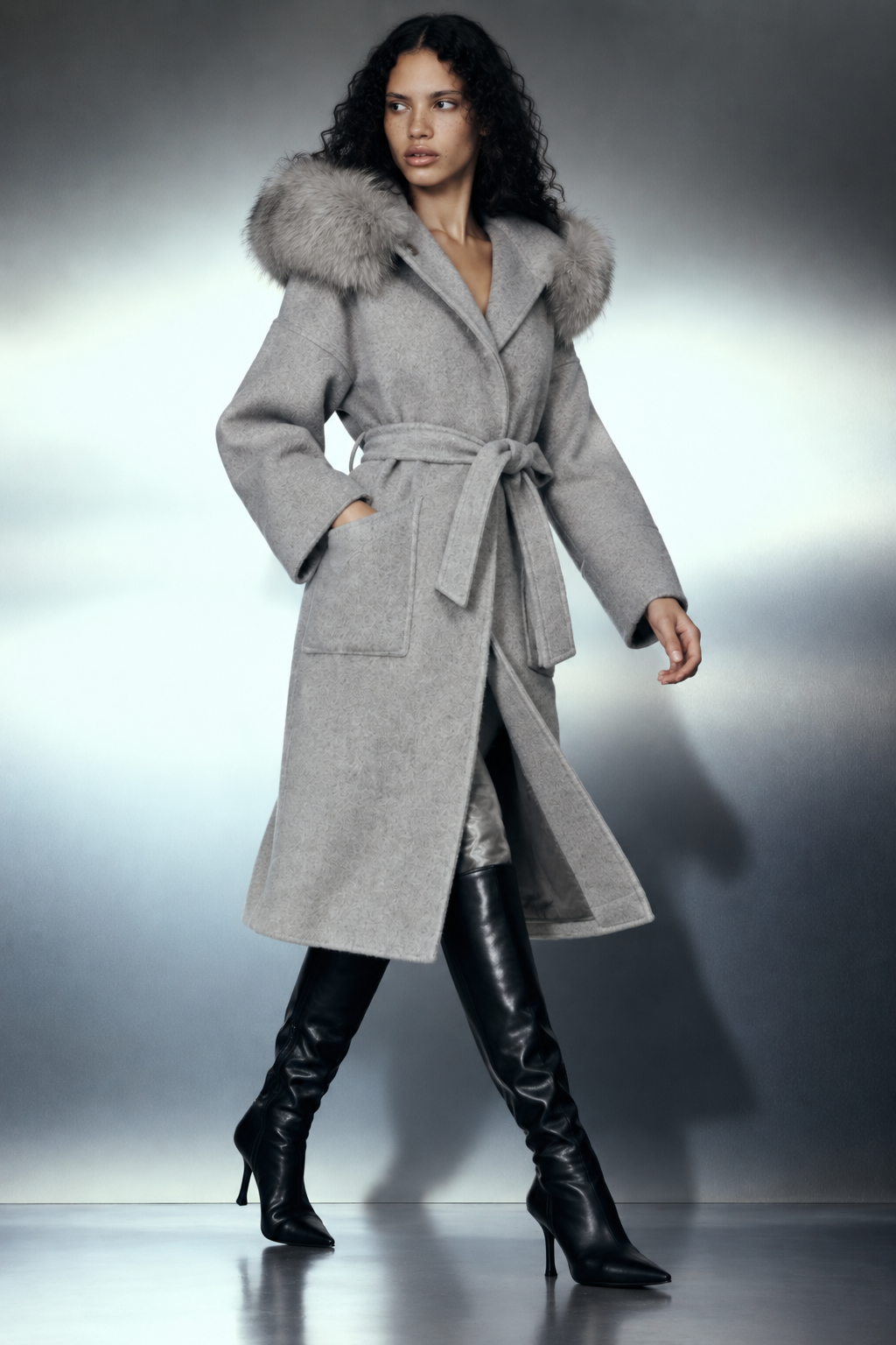 The Samara Long Coat — long manteau gris à capuche fourrure, porté de trois quarts en mouvement